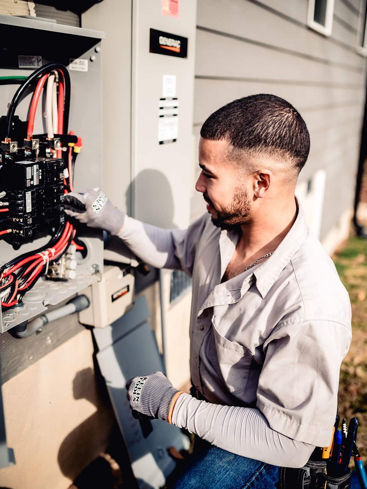 Smiling electrician in work gloves and tool belt standing beside a Generac transfer switch mounted on a home exterior