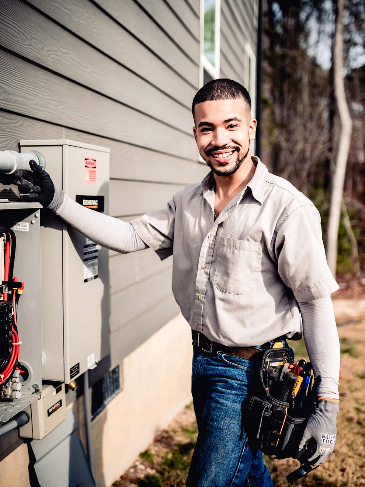Smiling electrician in work gloves and tool belt standing beside a Generac transfer switch mounted on a home exterior